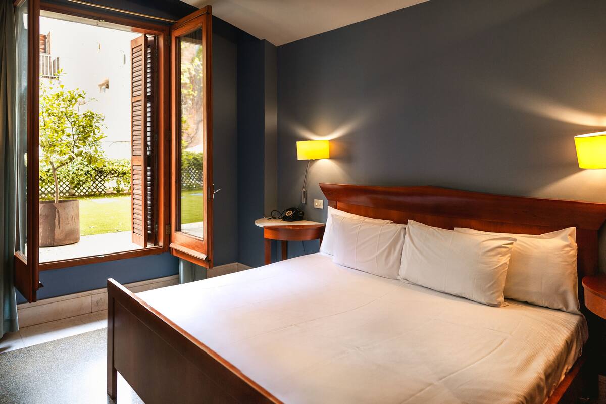 A bedroom with a bed, two bedside lamps, a window with open shutters, and a view of the outdoors.
