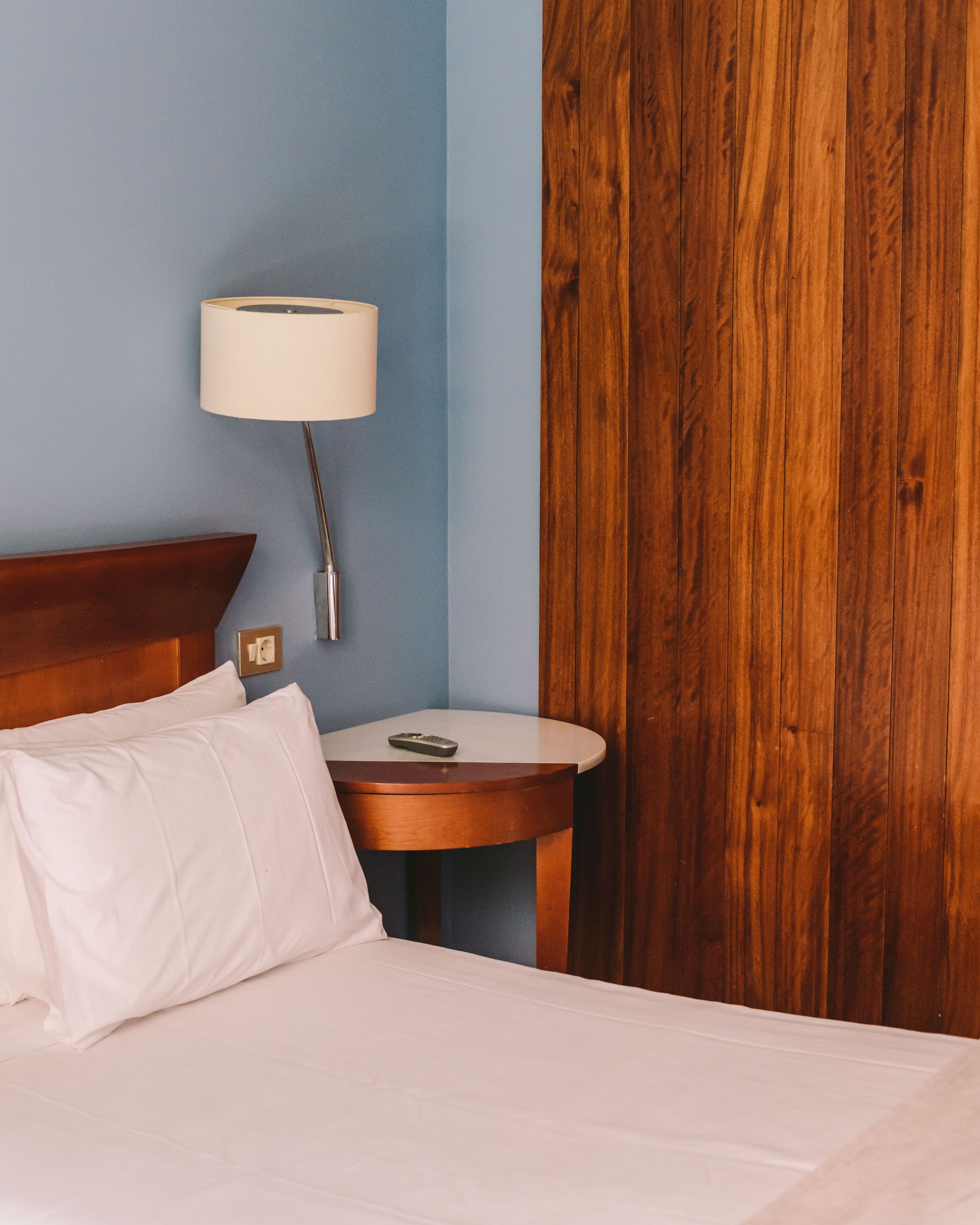 Una cama bien hecha con sábanas blancas, cabecera de madera, mesita de noche con lámpara y un panel de pared de madera.