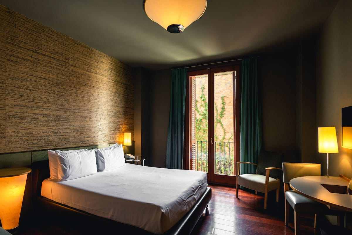 A hotel room with a large bed, two bedside tables with lamps, a small table with chairs, and a view of greenery through a sliding door.
