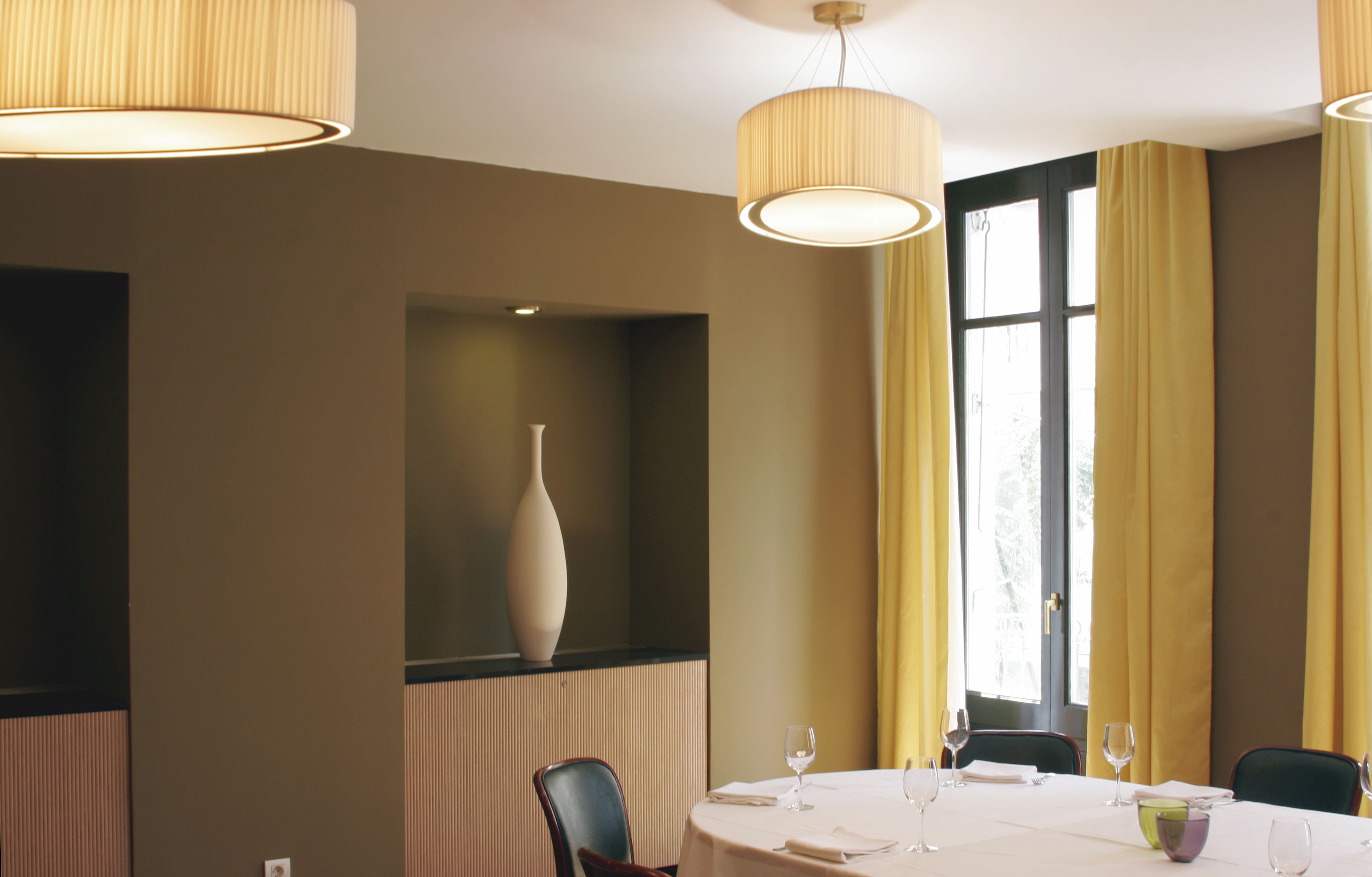 Comedor con una mesa redonda para cuatro personas, un florero en un estante y ventanas grandes con cortinas amarillas.