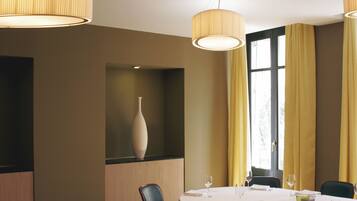 Comedor con una mesa redonda para cuatro personas, un florero en un estante y ventanas grandes con cortinas amarillas.
