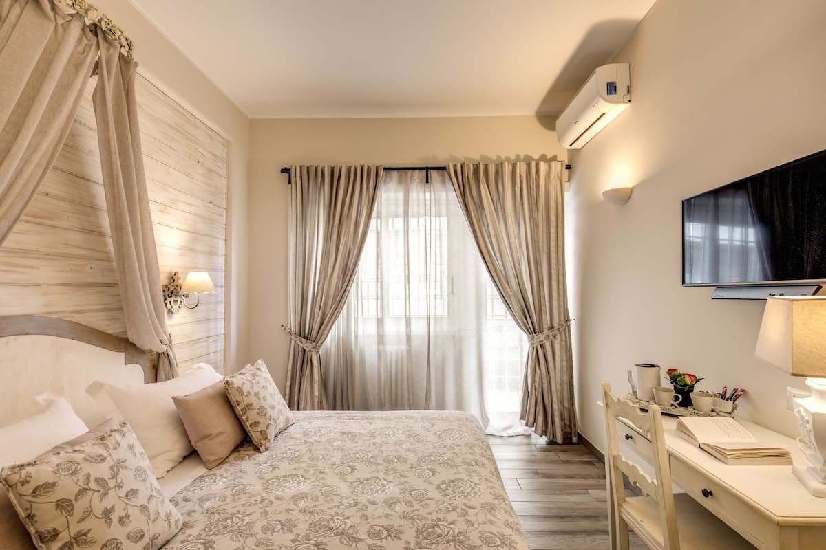 A bedroom with a bed, curtains, a desk, a TV, and an air conditioning unit.