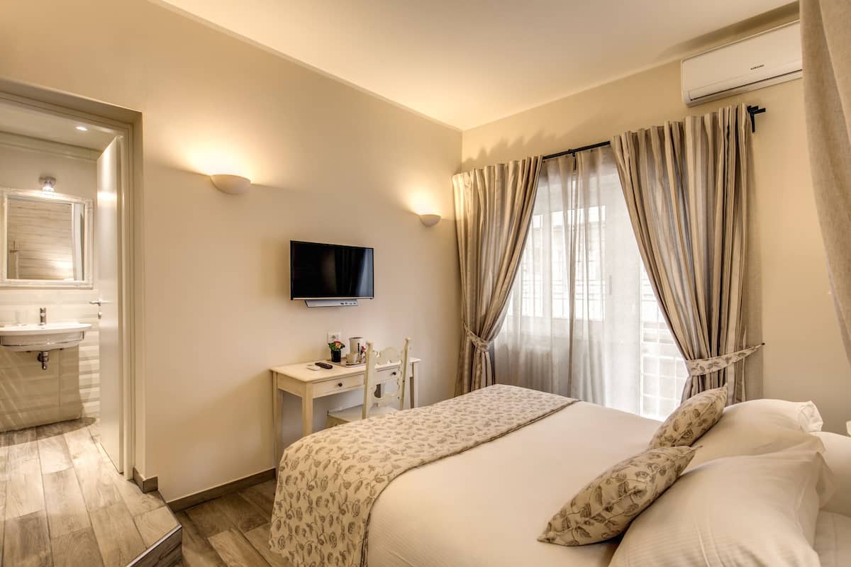 A hotel room with a bed, a TV, a desk with a chair, a vanity with a mirror, and a window with curtains.