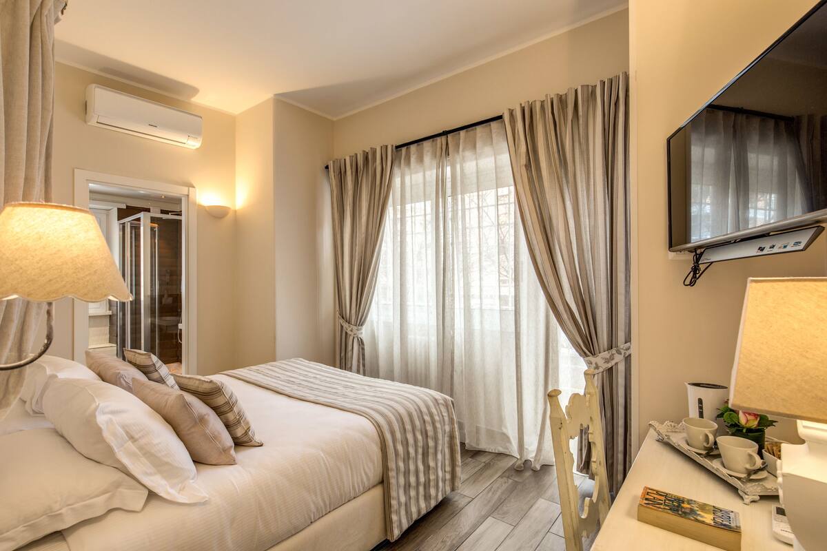 A hotel room with a bed, bedside lamps, a TV, a tray table with cups and a book, and a window with curtains.