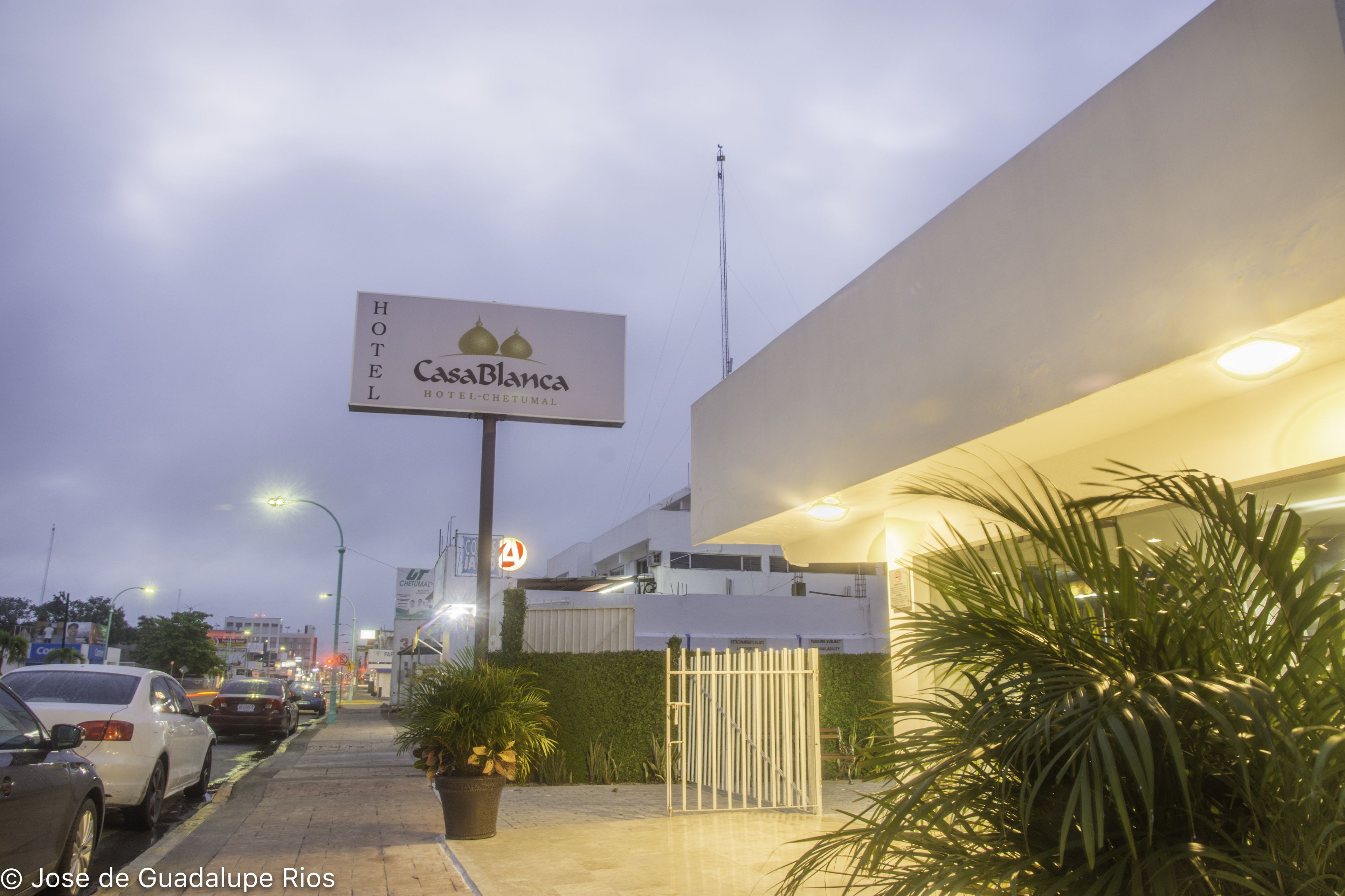 Une enseigne d’hôtel pour le Casa Blanca Hotel-Chetumal, un bâtiment moderne doté d’une entrée sécurisée par un portail, de voitures garées et d’un lampadaire.