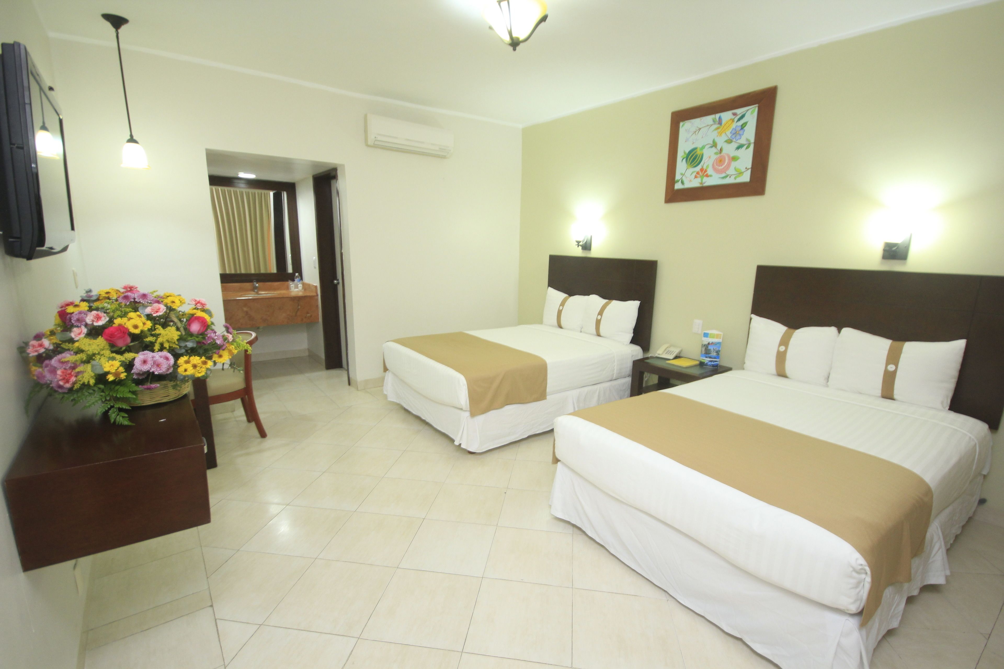 A hotel room with two beds, a desk with a flower arrangement, a mirror, and a painting on the wall.