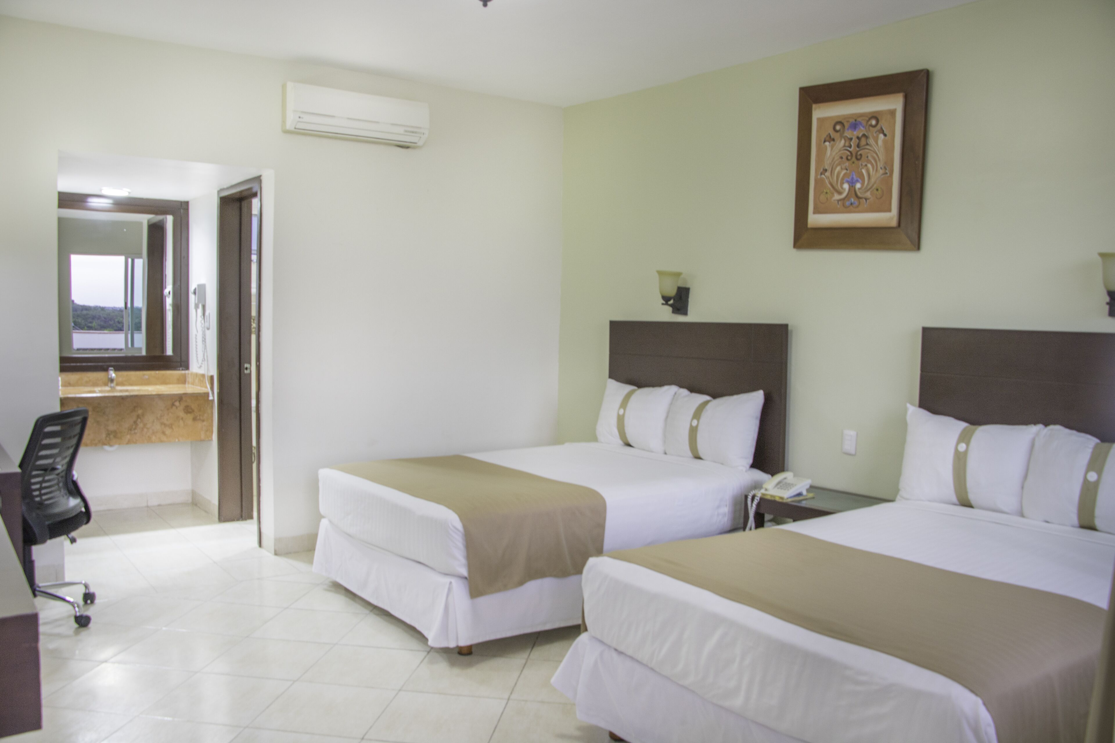 Habitación de hotel con dos camas, un escritorio y un baño visible a través de una puerta abierta.