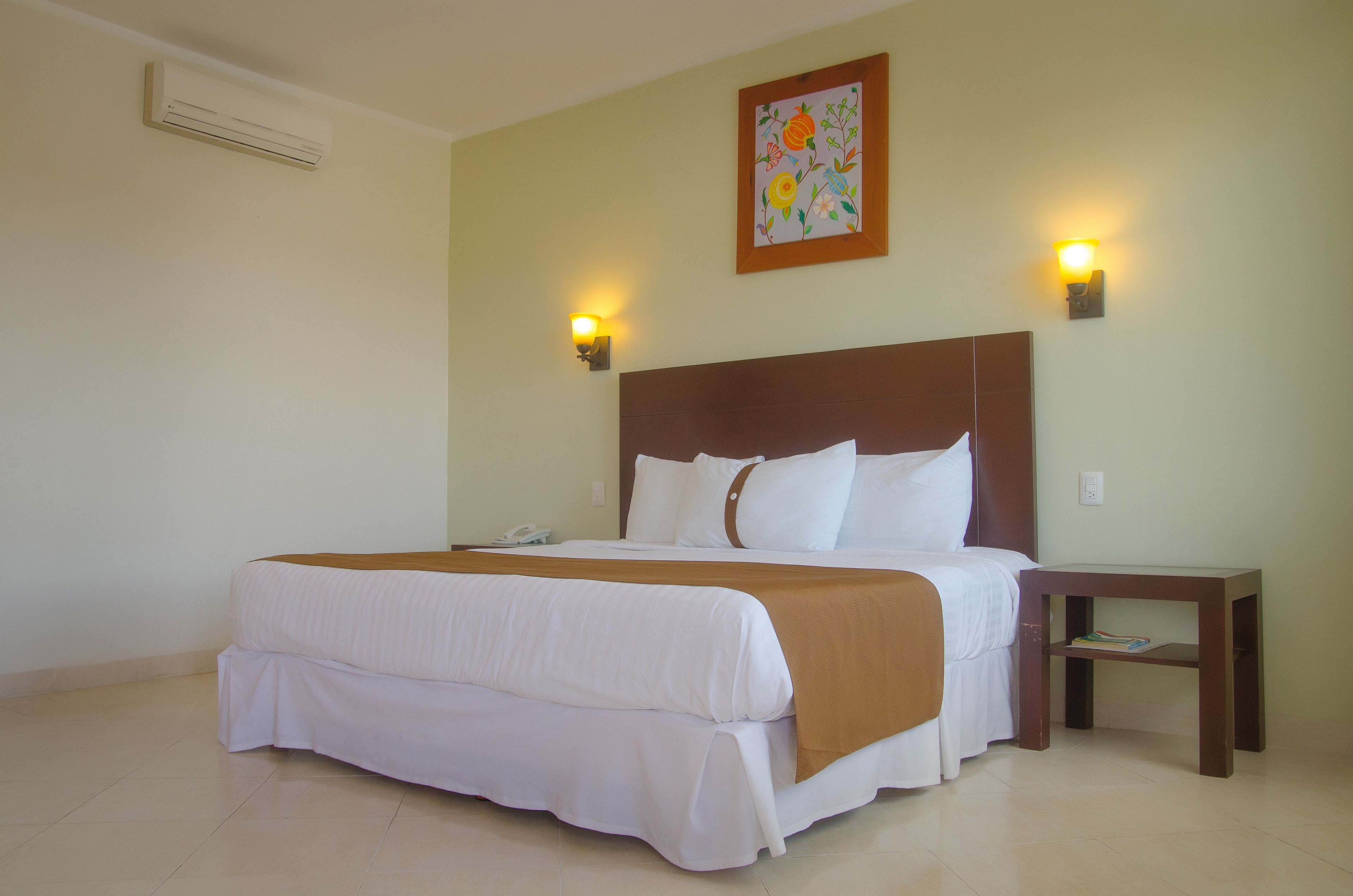 A neatly made bed with white linens and a brown throw, a wooden nightstand with a lamp, and a floral painting on the wall.