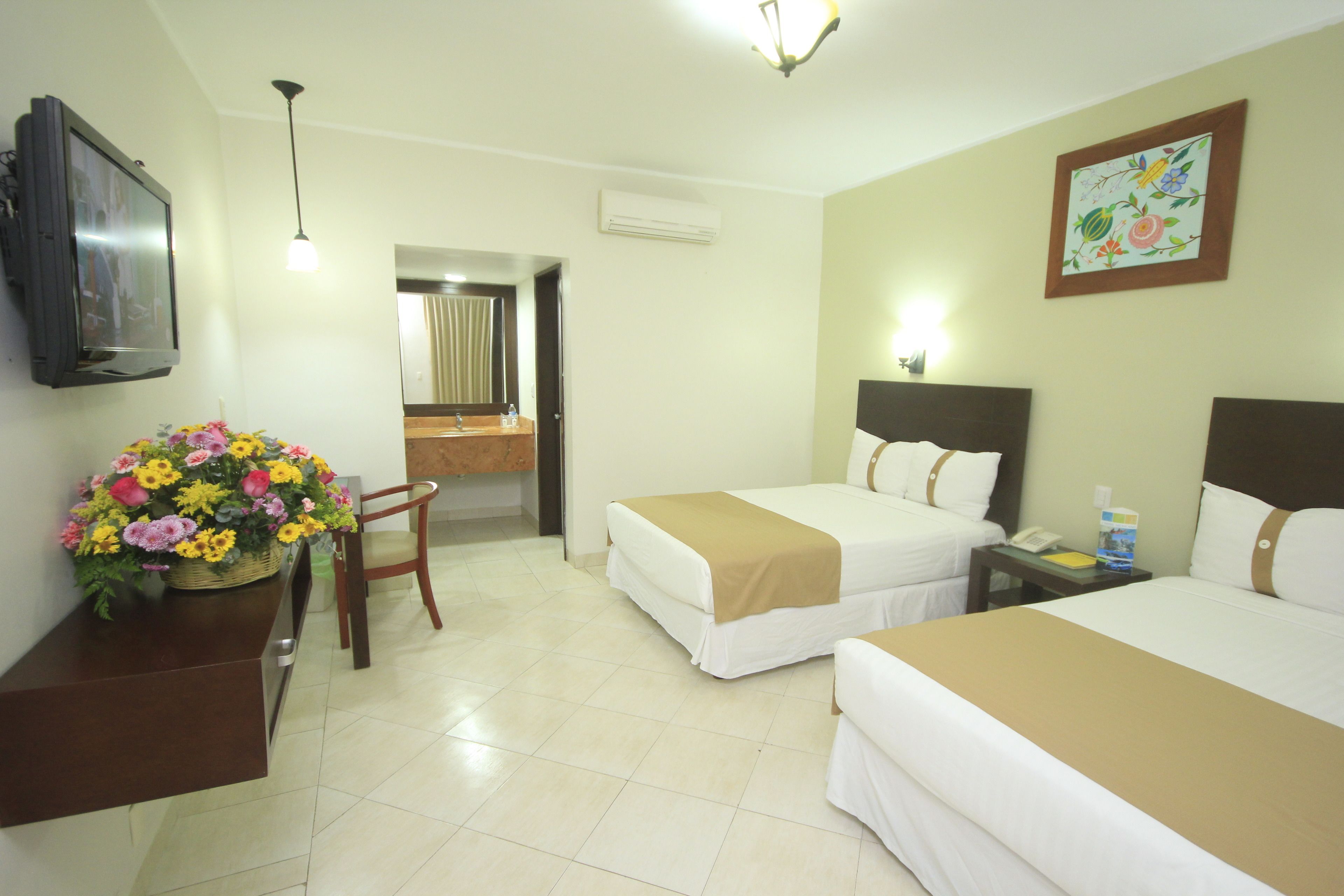Habitación de hotel con dos camas, un escritorio con televisor, una silla, un baño y una decoración floral.