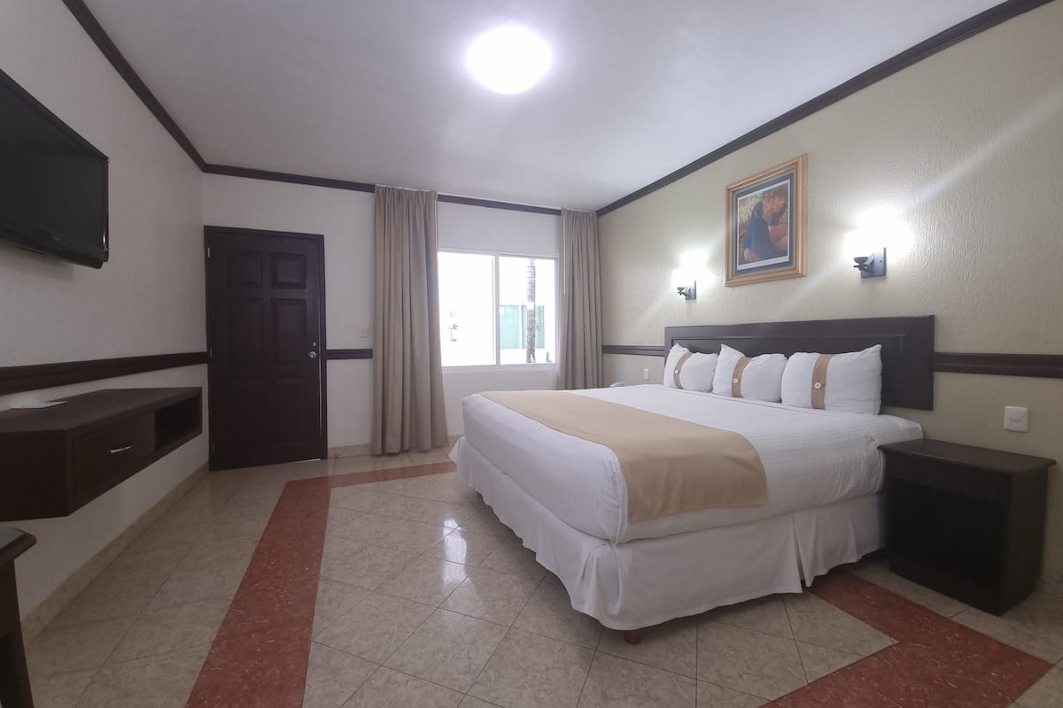 Habitación de hotel con cama, mesita de noche, televisor, ventana con cortinas y un cuadro en la pared.