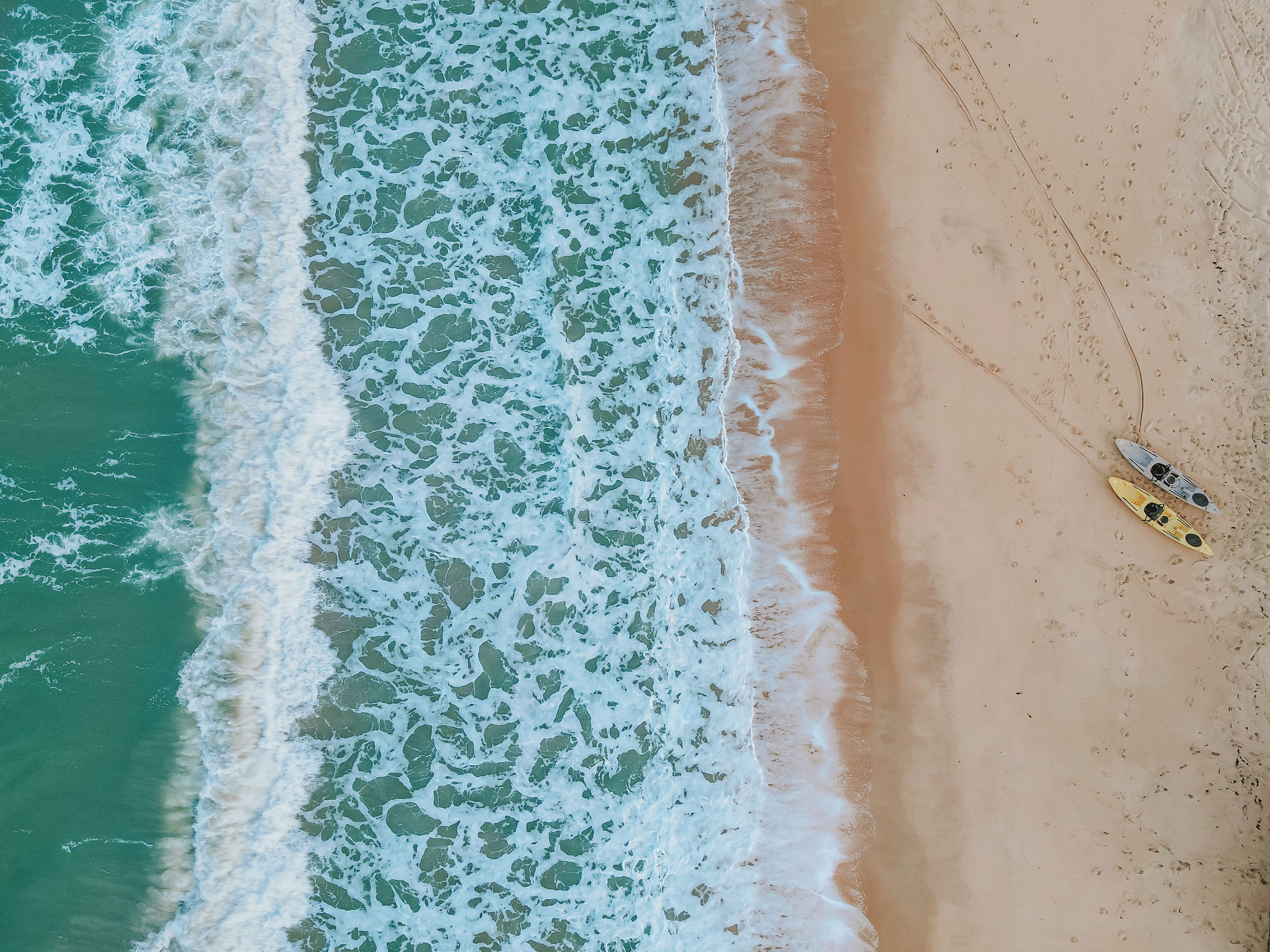An aerial view of a beach with waves crashing onto the shore and two kayaks on the sand.