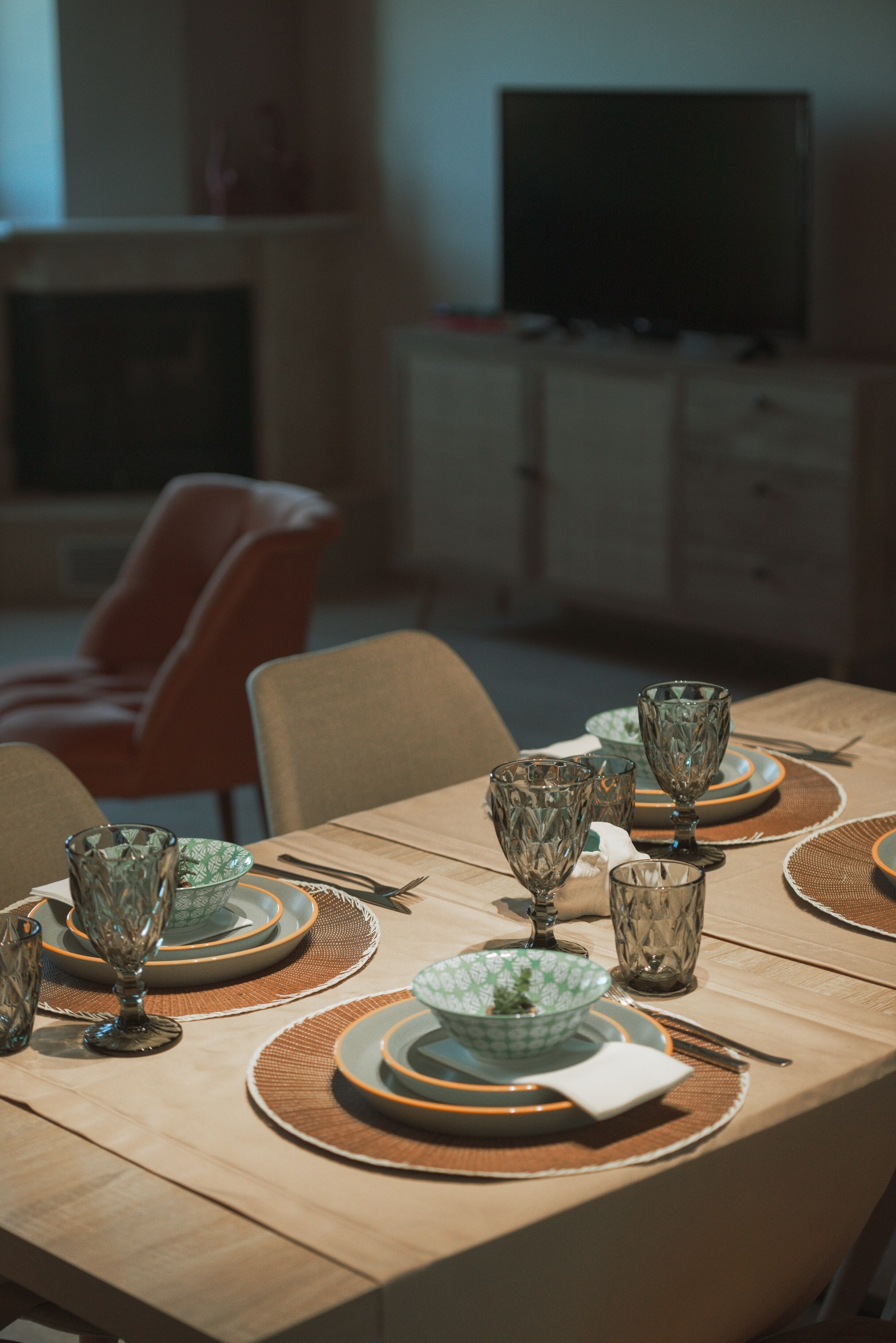 Una mesa de comedor dispuesta con platos, tazones y vasos, con un televisor y una silla roja al fondo.