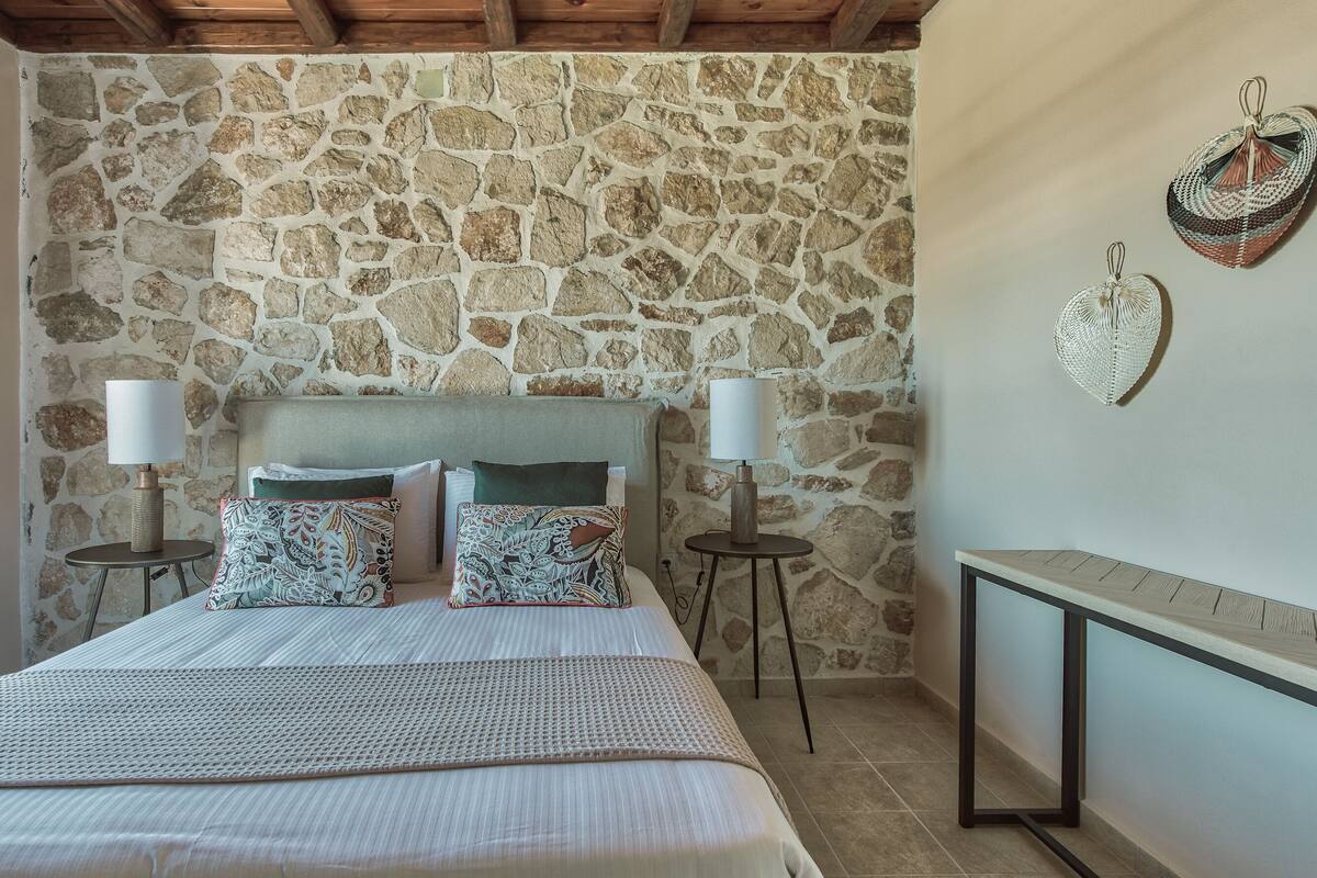 A bedroom with a stone wall, a bed with patterned pillows, two bedside lamps, and a woven headboard.