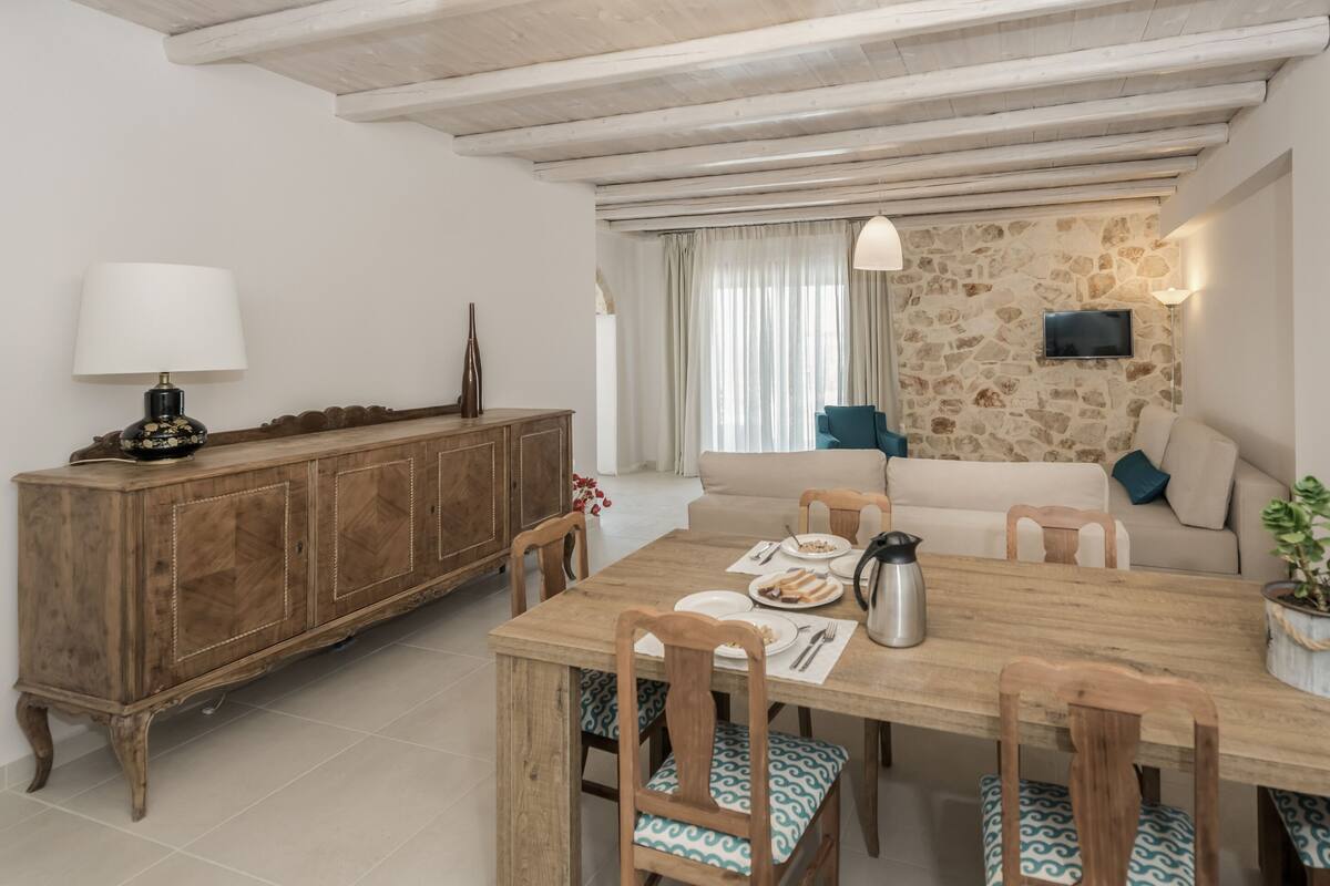 A dining area with a wooden table, chairs, a cabinet, a sofa, and a television mounted on a stone wall.