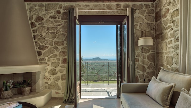 A cozy living room with a stone wall, a sofa, a coffee table with a teapot, and a view of a mountainous landscape through an open door. - Agrikia Villa (Zakynthos)