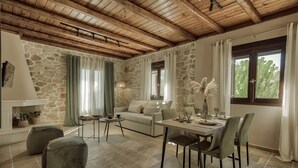 A cozy living room with a stone wall, wooden ceiling beams, a sofa, a dining table with chairs, and a fireplace. - Agrikia Villa (Zakynthos)