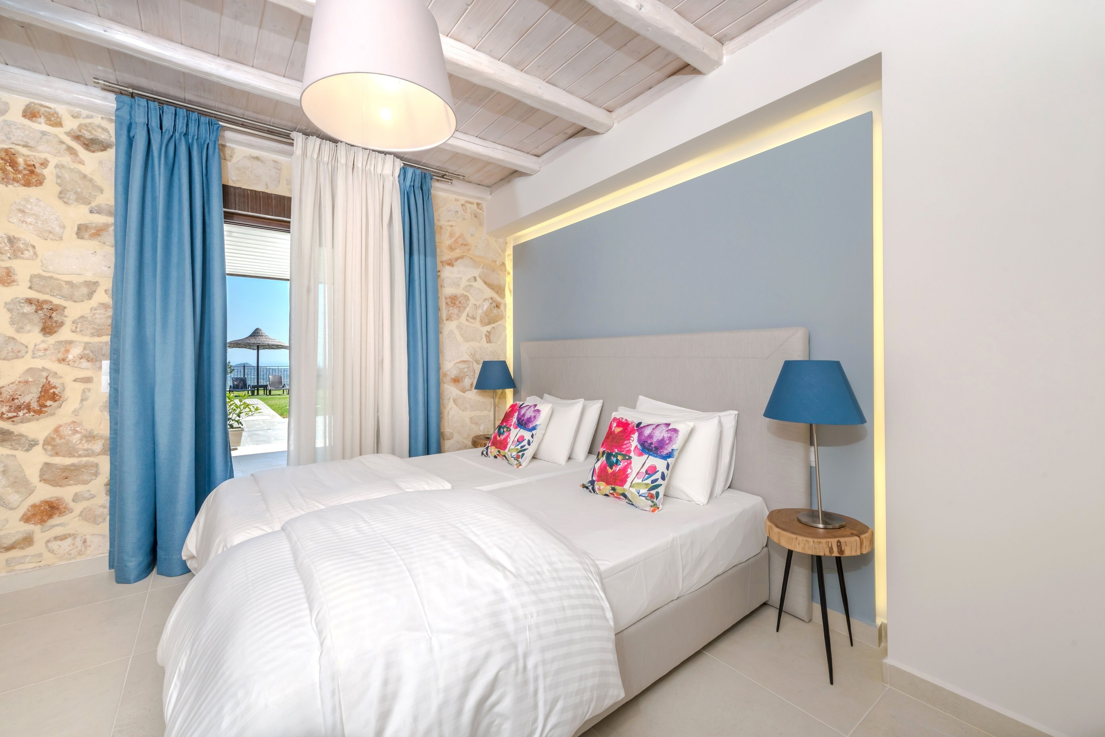 Un dormitorio con pared de piedra, una cama grande con ropa de cama blanca, dos lámparas y vistas al exterior.