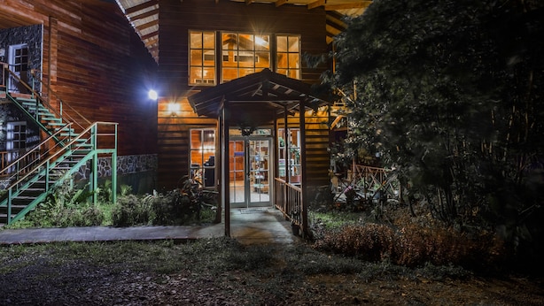 A wooden cabin with a staircase, a covered entrance, and a well-lit interior visible through the windows. - Hotel Bosque Verde Lodge (Sierra)