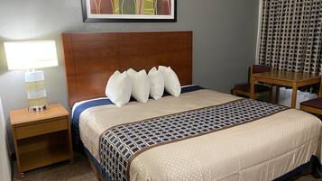 A hotel room with a wooden headboard, a bed with a patterned bedspread, a nightstand with a lamp, a framed artwork above the bed, and a small wooden table with chairs.