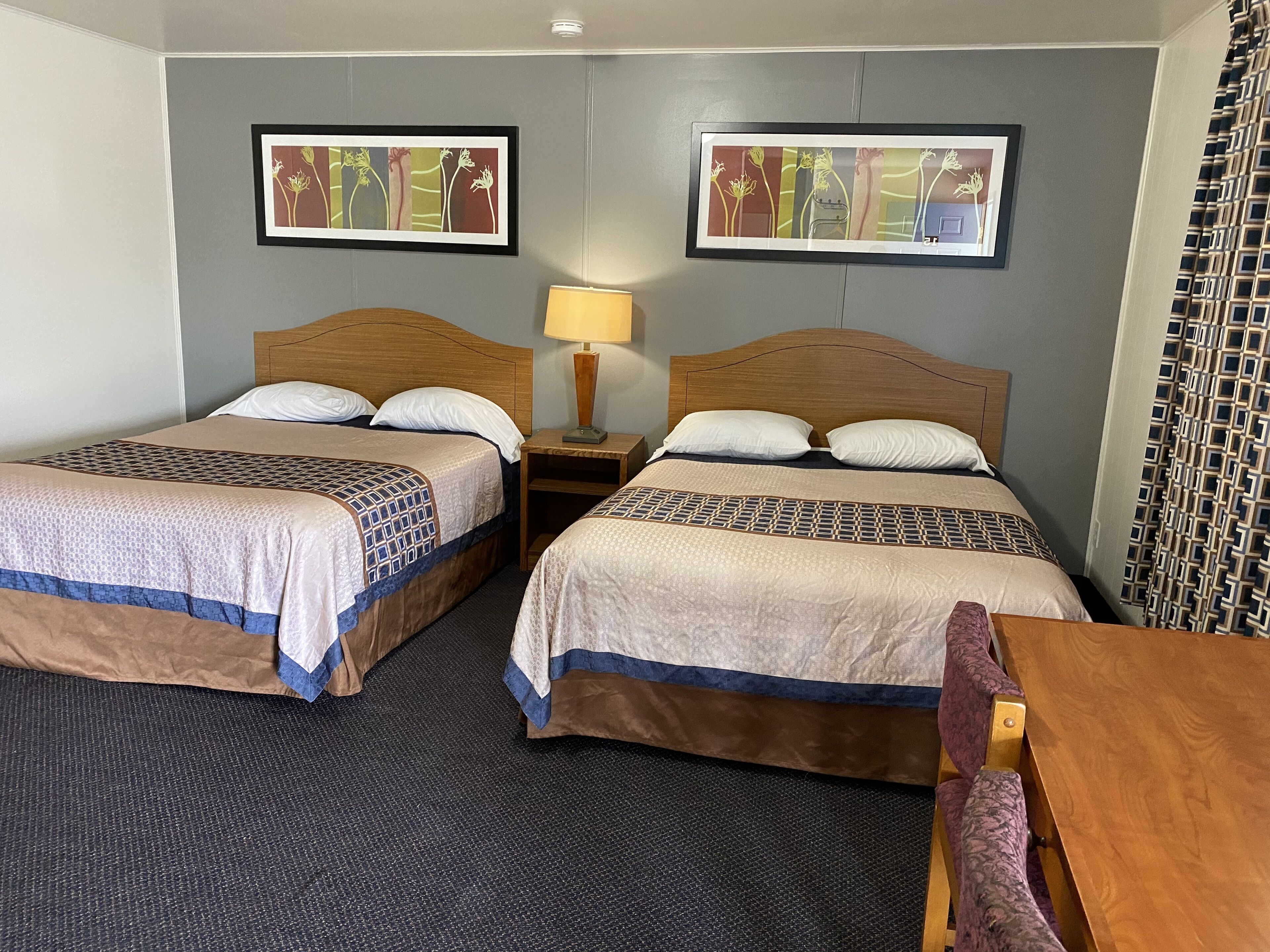 A hotel room with two beds, a wooden headboard, a nightstand with a lamp, and a framed picture on the wall.