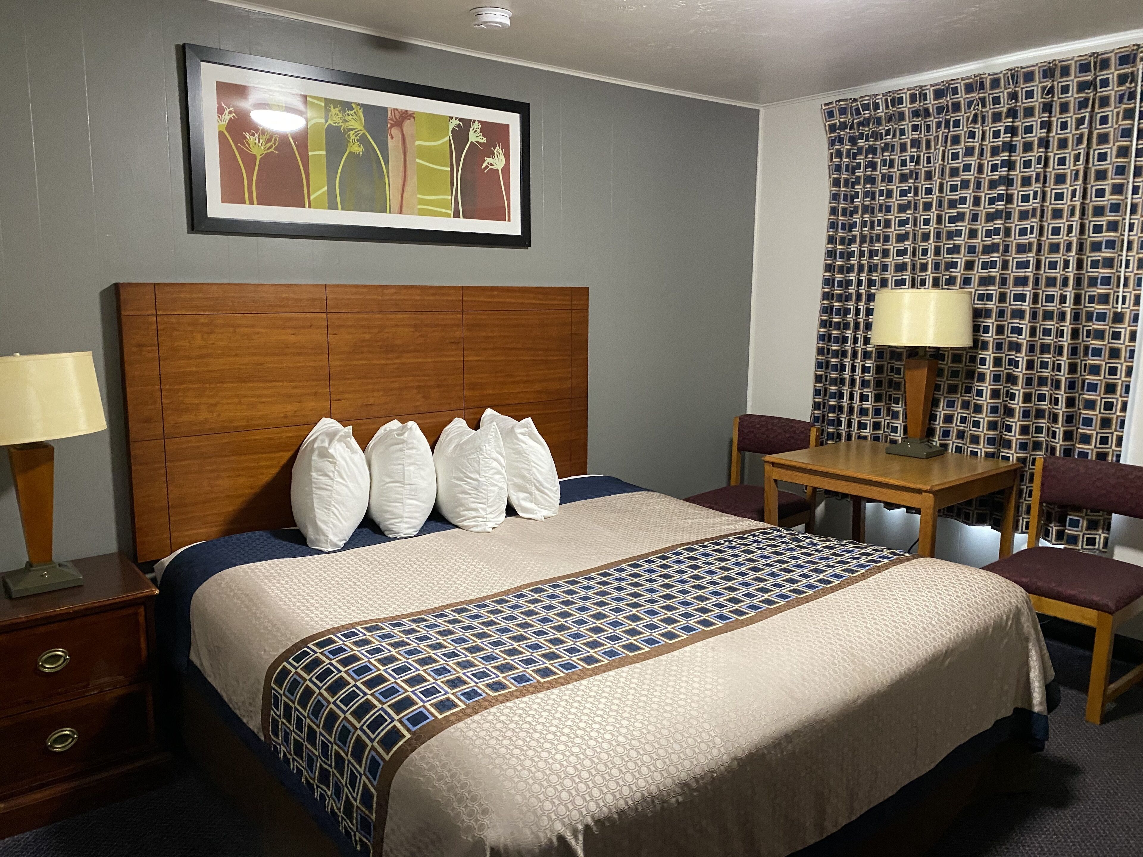 A hotel room with a large bed, a wooden headboard, a desk with a chair, a lamp, and a patterned wall.