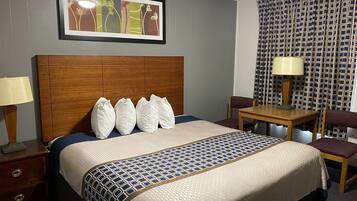 A hotel room with a large bed, a wooden headboard, a desk with a chair, a lamp, and a patterned wall.