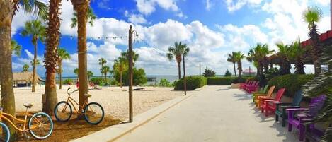 A beachside walkway with bicycles, colorful chairs, and palm trees.
