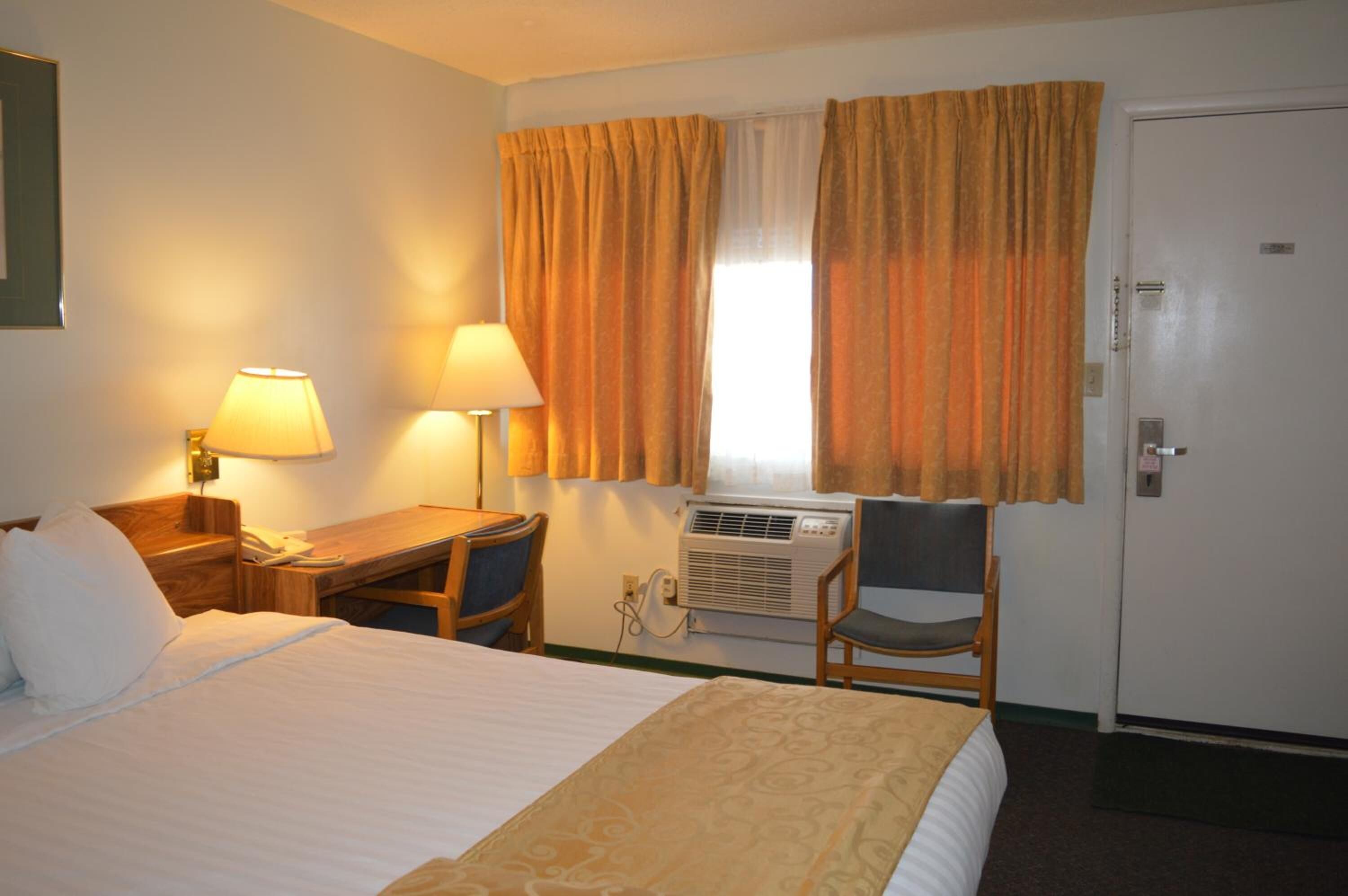 A hotel room with a bed, two bedside lamps, a desk with a chair, an air conditioning unit, and a door.