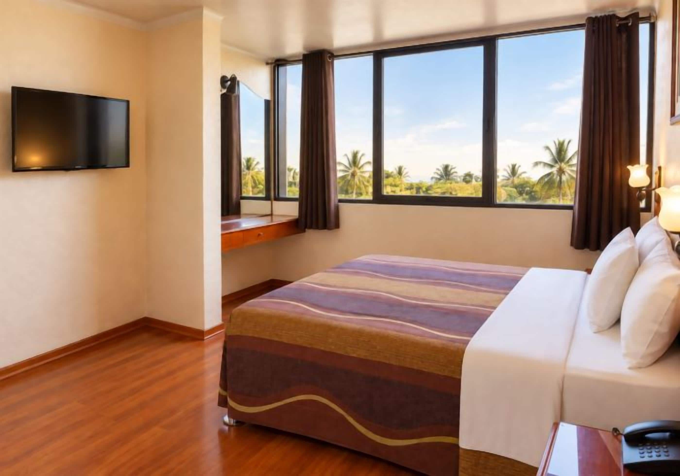 A hotel room with a bed, a television, a window with curtains, and a view of palm trees.