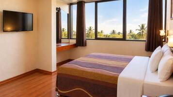 A hotel room with a bed, a television, a window with curtains, and a view of palm trees.