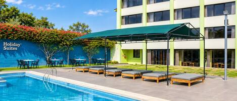 A hotel pool area with lounge chairs and tables, a covered seating area, and a building with a sign reading 'Holiday Suites'.