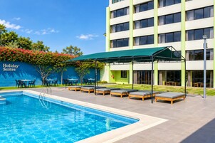 Zona de piscina hotelera con sillones de descanso y mesas, un área de estar cubierta y un edificio con un letrero que dice 'Holiday Suites'.
