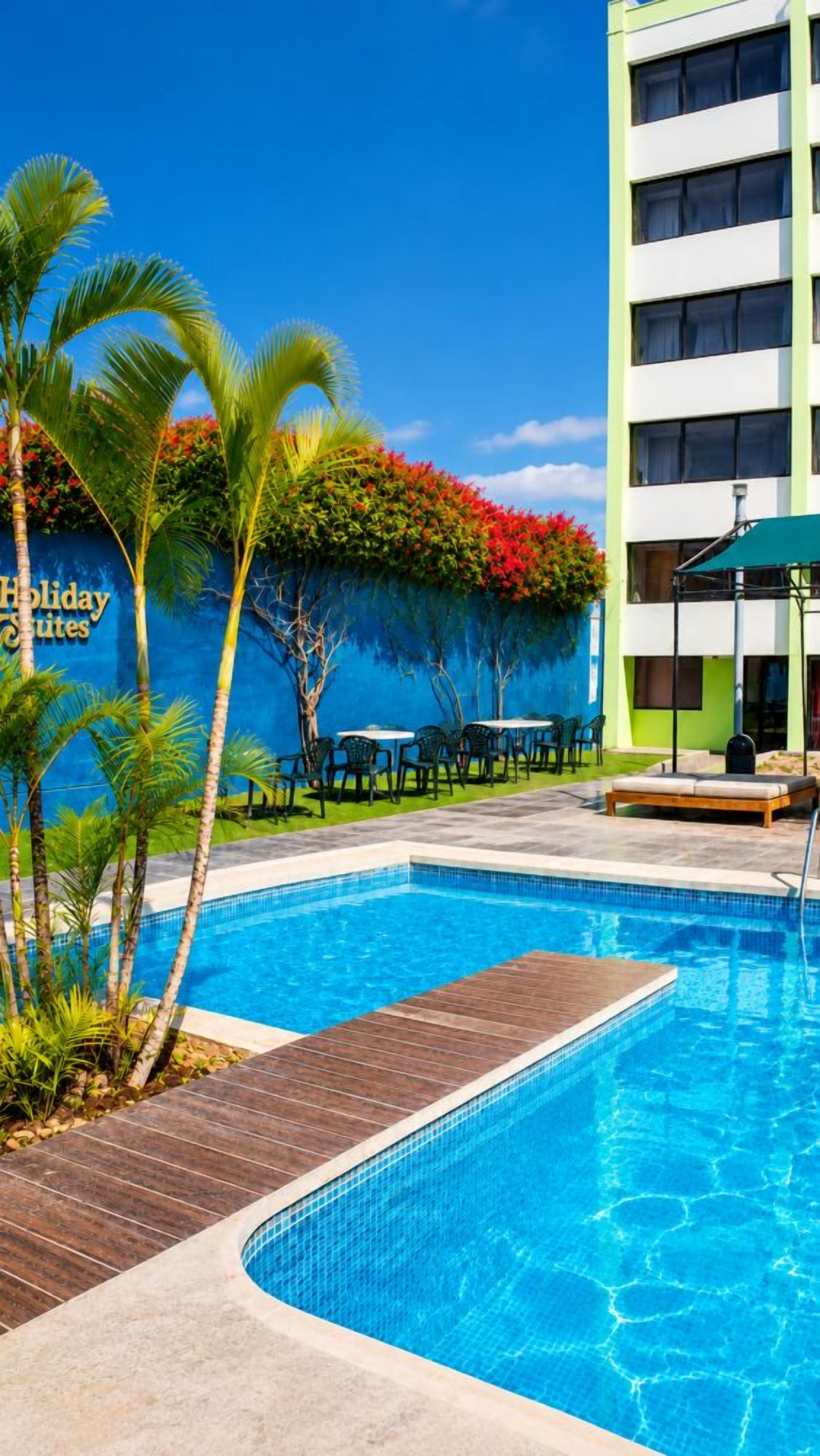 A swimming pool with a wooden deck, surrounded by palm trees and a building with a sign that reads 'Holiday Suites'.