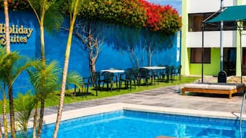 A swimming pool with a wooden deck, surrounded by palm trees and a building with a sign that reads 'Holiday Suites'.