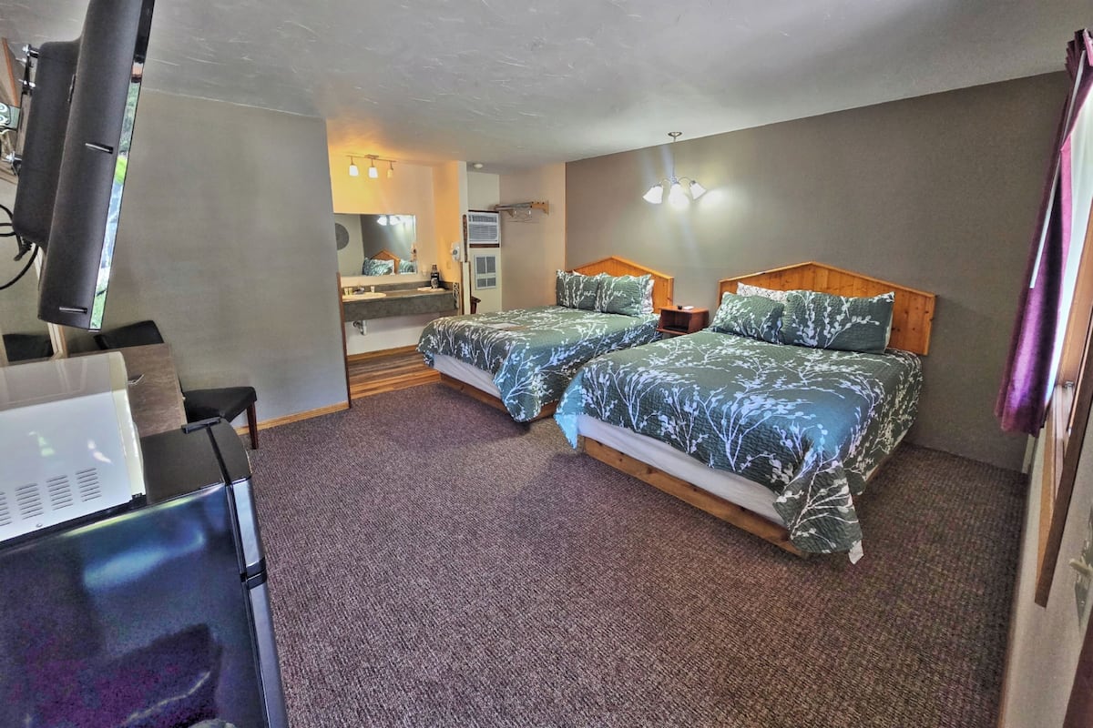 A hotel room with two beds, a desk with a computer, a mirror, and a bathroom with a sink and mirror.