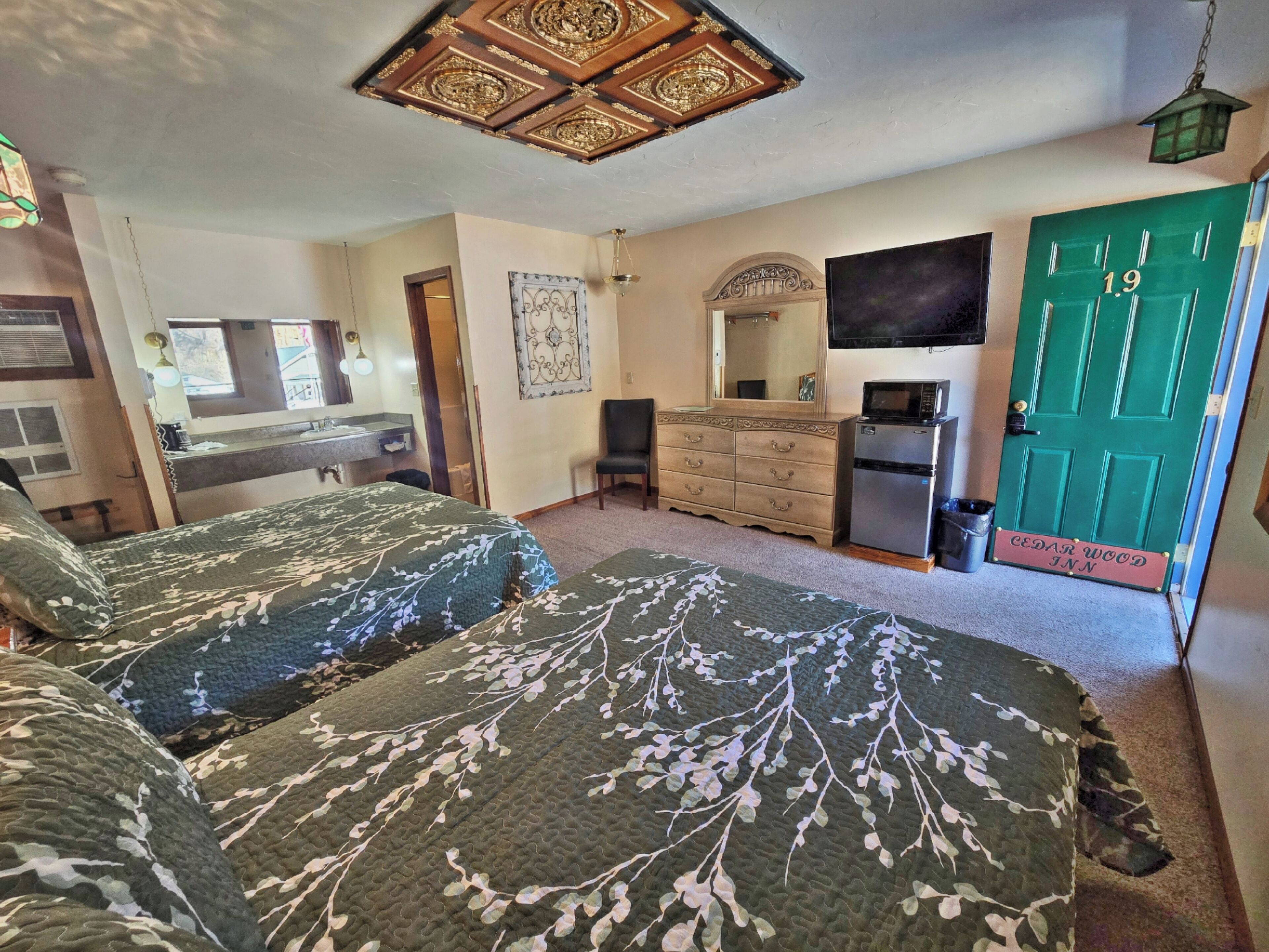 Habitación con dos camas, un aparador, un espejo, un televisor y una puerta verde con la inscripción 'Cedar Wood Inn'.