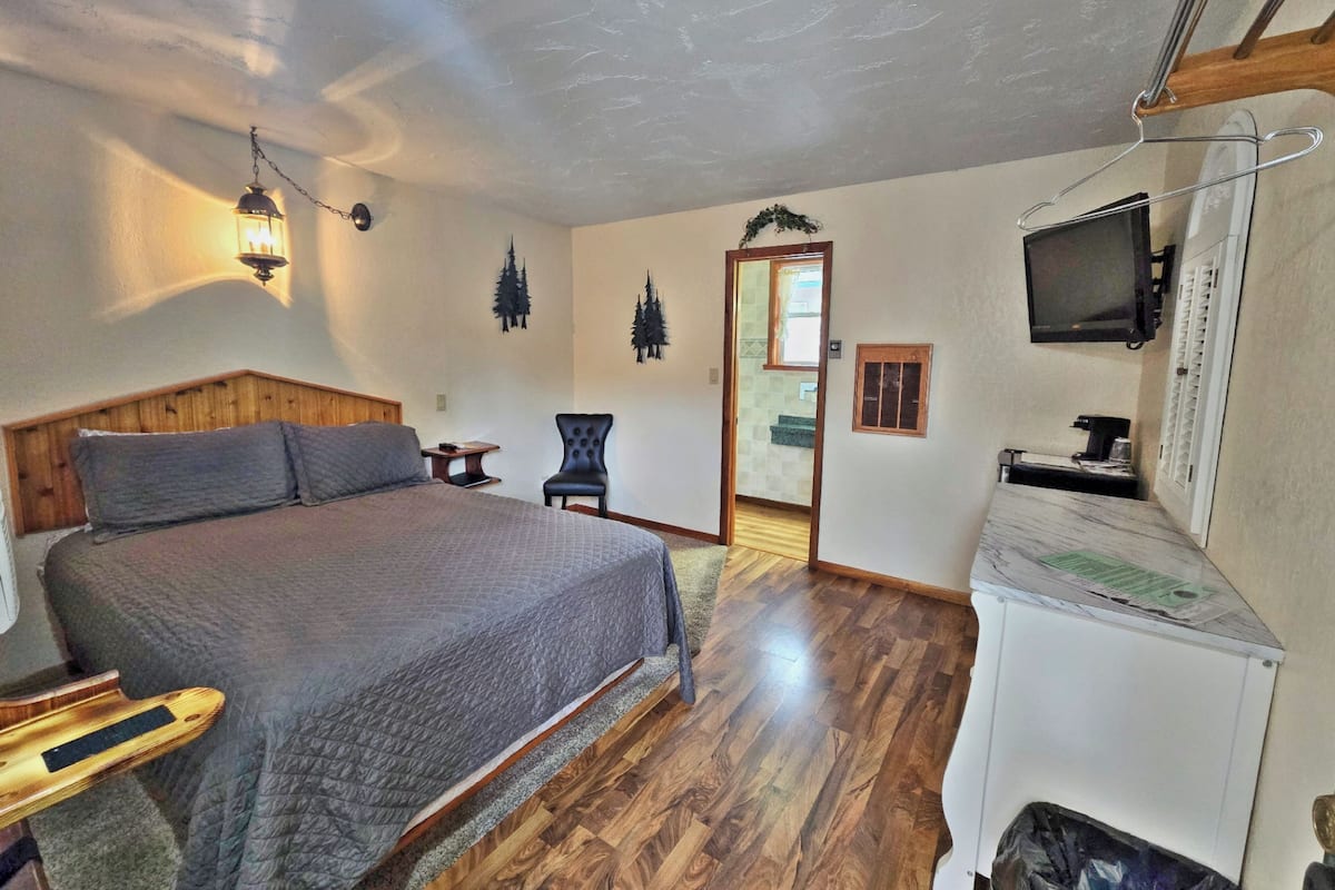 A bedroom with a wooden headboard, a bed with a gray quilt, a small table, a chair, a TV mounted on the wall, and a door leading to another room.
