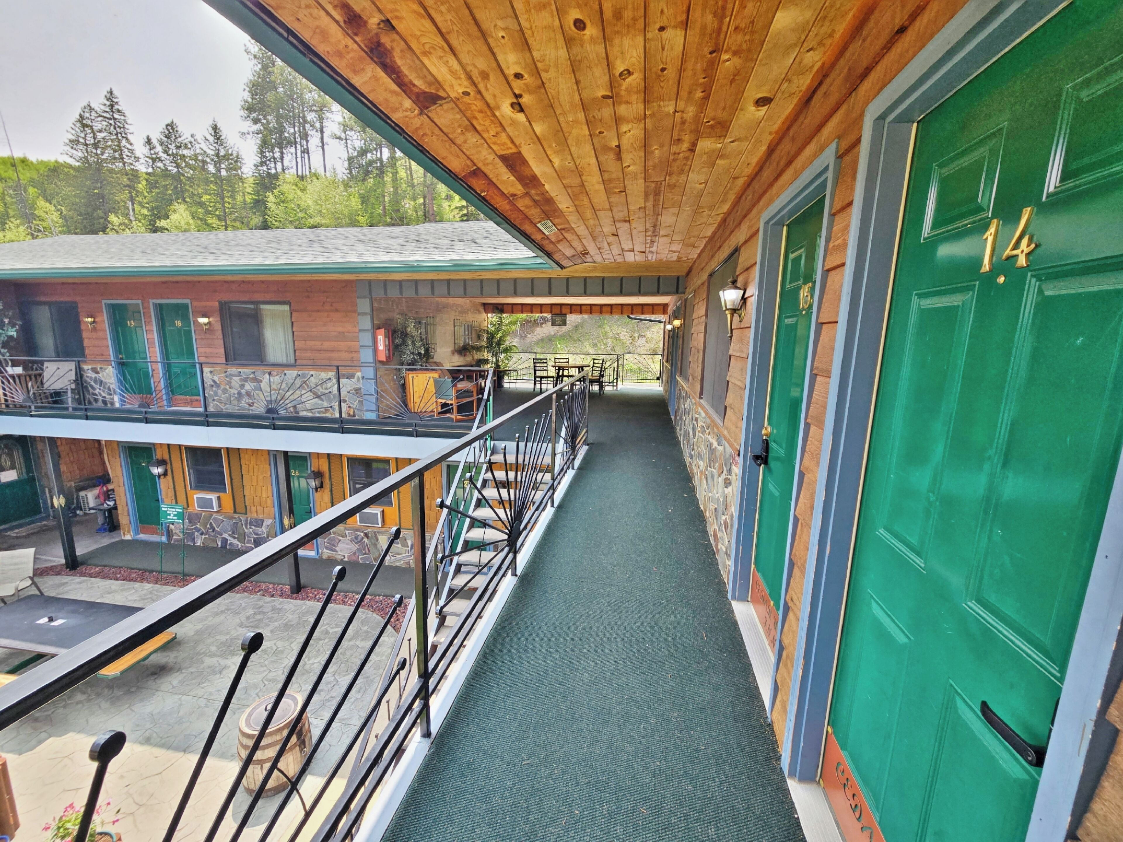 A covered outdoor corridor with a wooden ceiling, green doors, and a view of a building with multiple windows and a staircase.