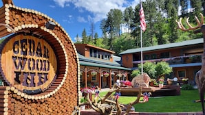 A lodge with a circular wooden sign reading "Cedar Wood Inn" surrounded by a landscaped area with a large tree sculpture and a flagpole with an American flag. - Cedar Wood Inn (Deadwood)