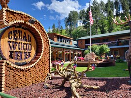 Una posada con un letrero de madera circular que dice "Cedar Wood Inn", rodeada de un área ajardinada con una gran escultura de árbol y un asta de bandera con la bandera estadounidense.