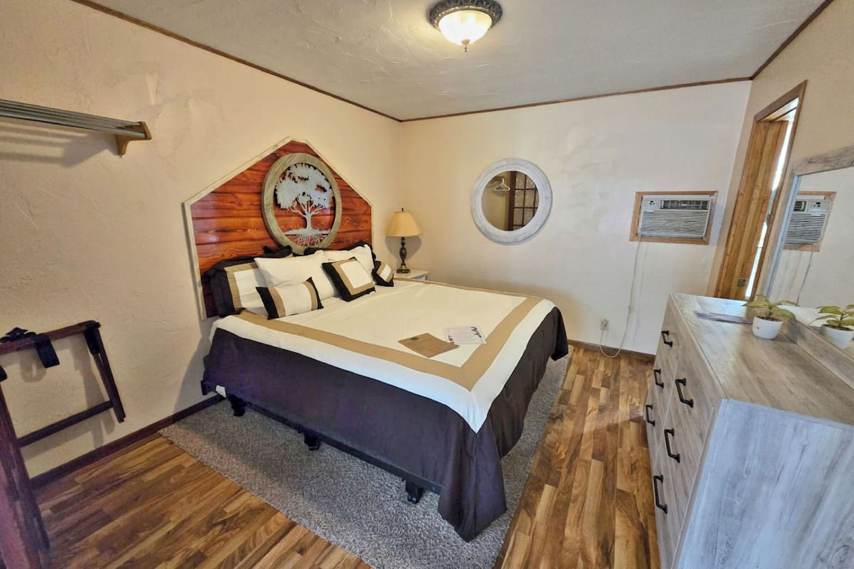 A bedroom with a wooden headboard, a bed with a patterned bedspread, a round mirror, and a wooden dresser with plants.