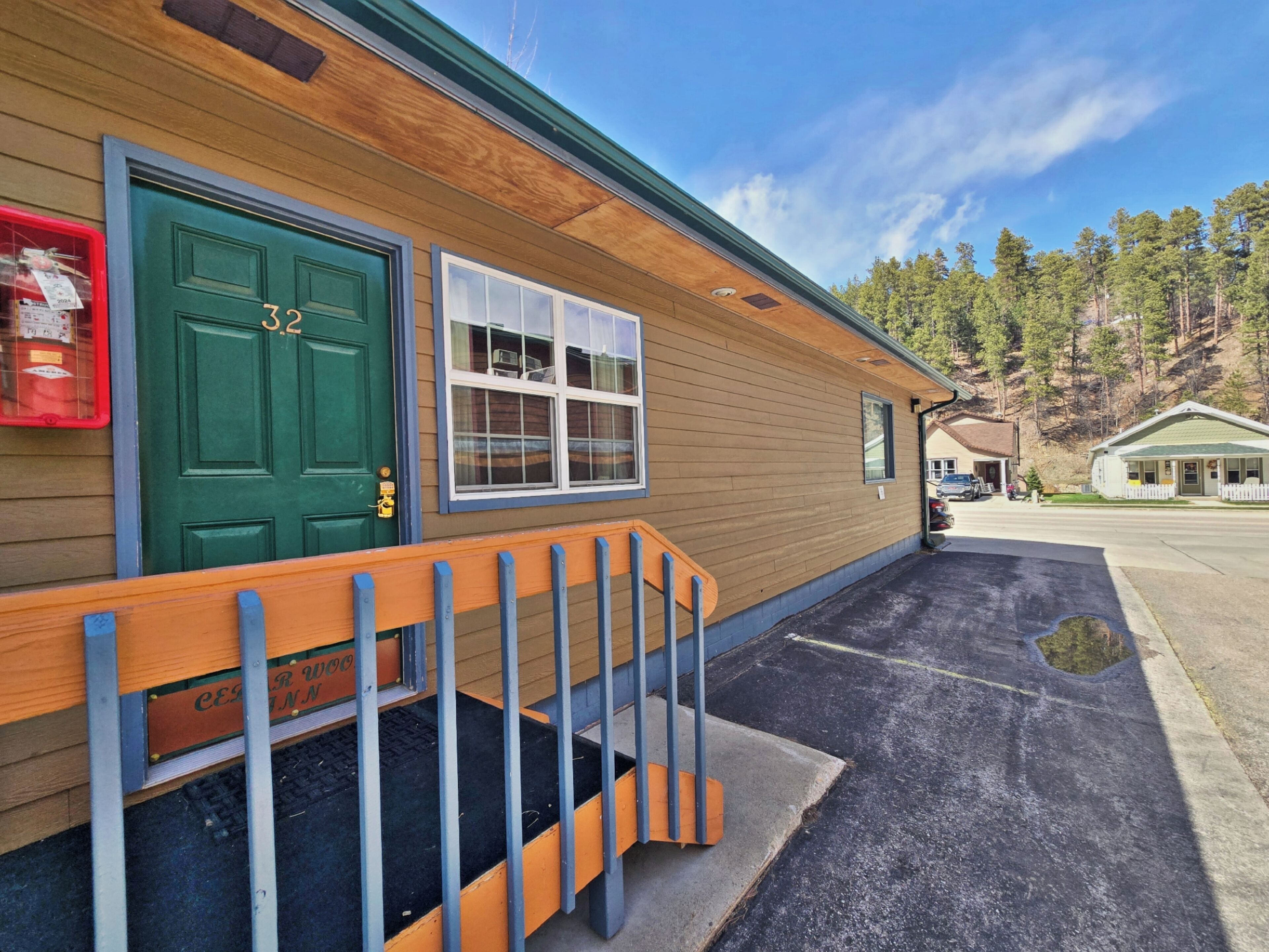 A wooden building with a green door and windows, a wooden porch with a metal railing, and a parking lot in front.