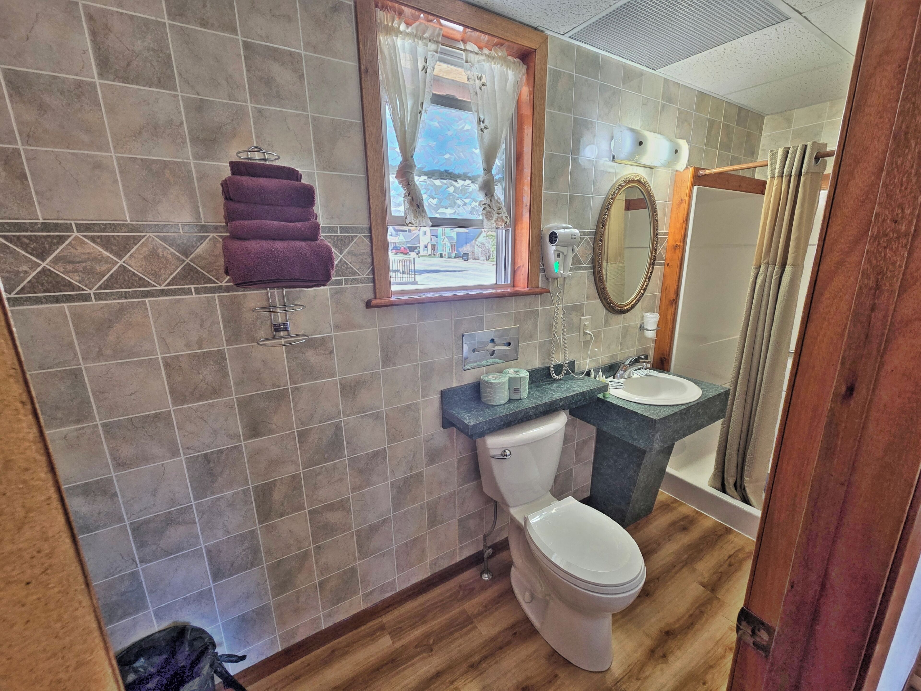 A bathroom with a toilet, sink, and shower. There is a window with curtains, a towel rack, and a mirror.