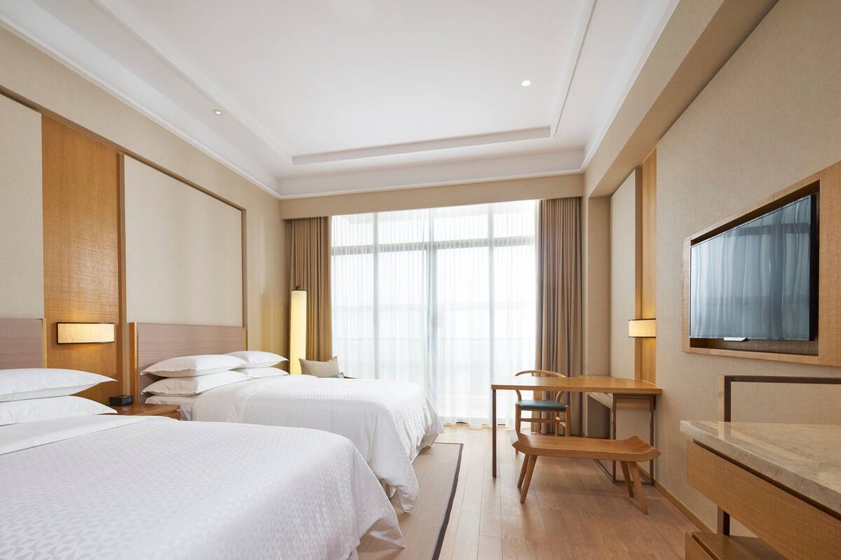 Quarto de hotel com duas camas, uma escrivaninha, uma cadeira, televisão e janelas amplas.