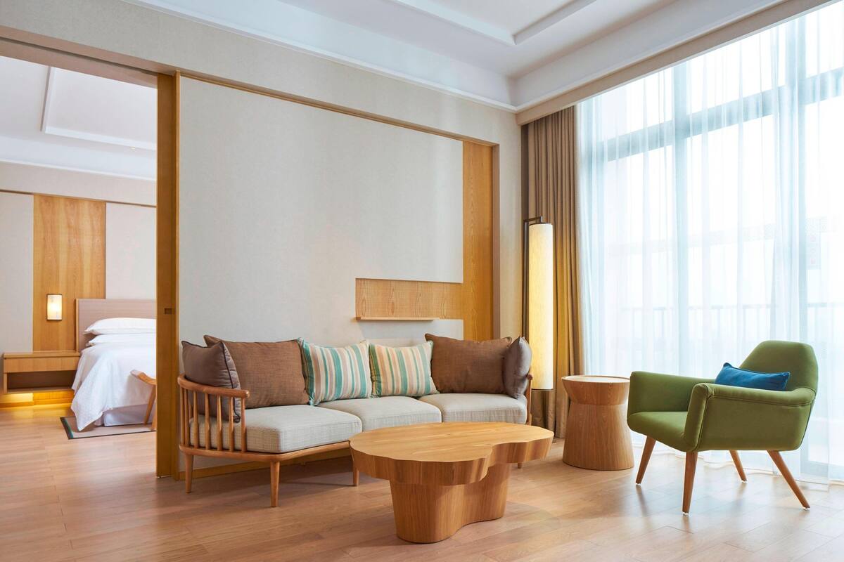 Quarto de hotel moderno com piso de madeira, um sofá, uma poltrona verde, mesa de centro de madeira e uma janela grande com cortinas.