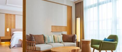 Una habitación de hotel moderna con piso de madera, un conjunto de sofás, un sillón verde, una mesa de centro de madera y un ventanal con cortinas.