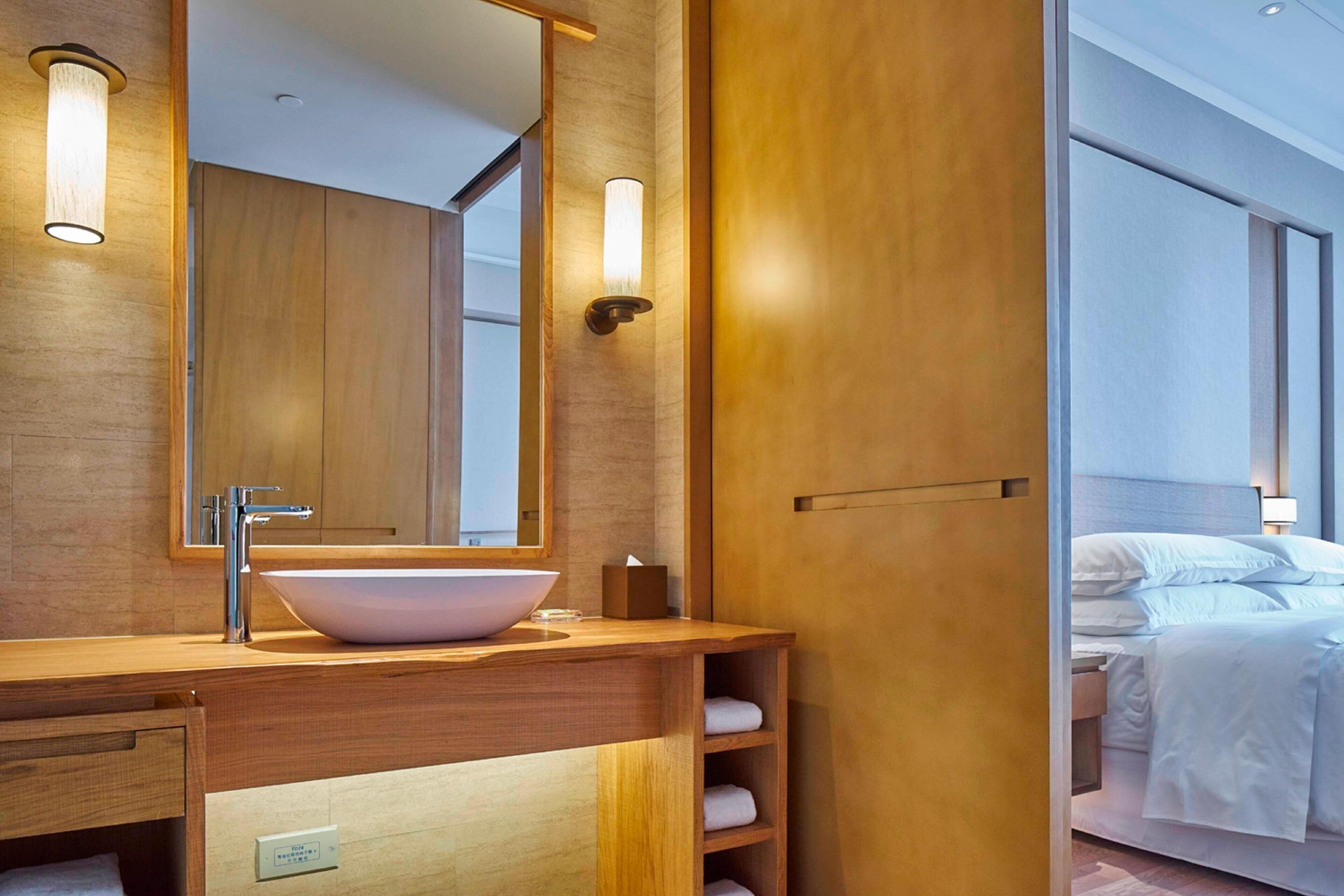 A modern bathroom with a wooden vanity, a large mirror, and a bedside table with a lamp.