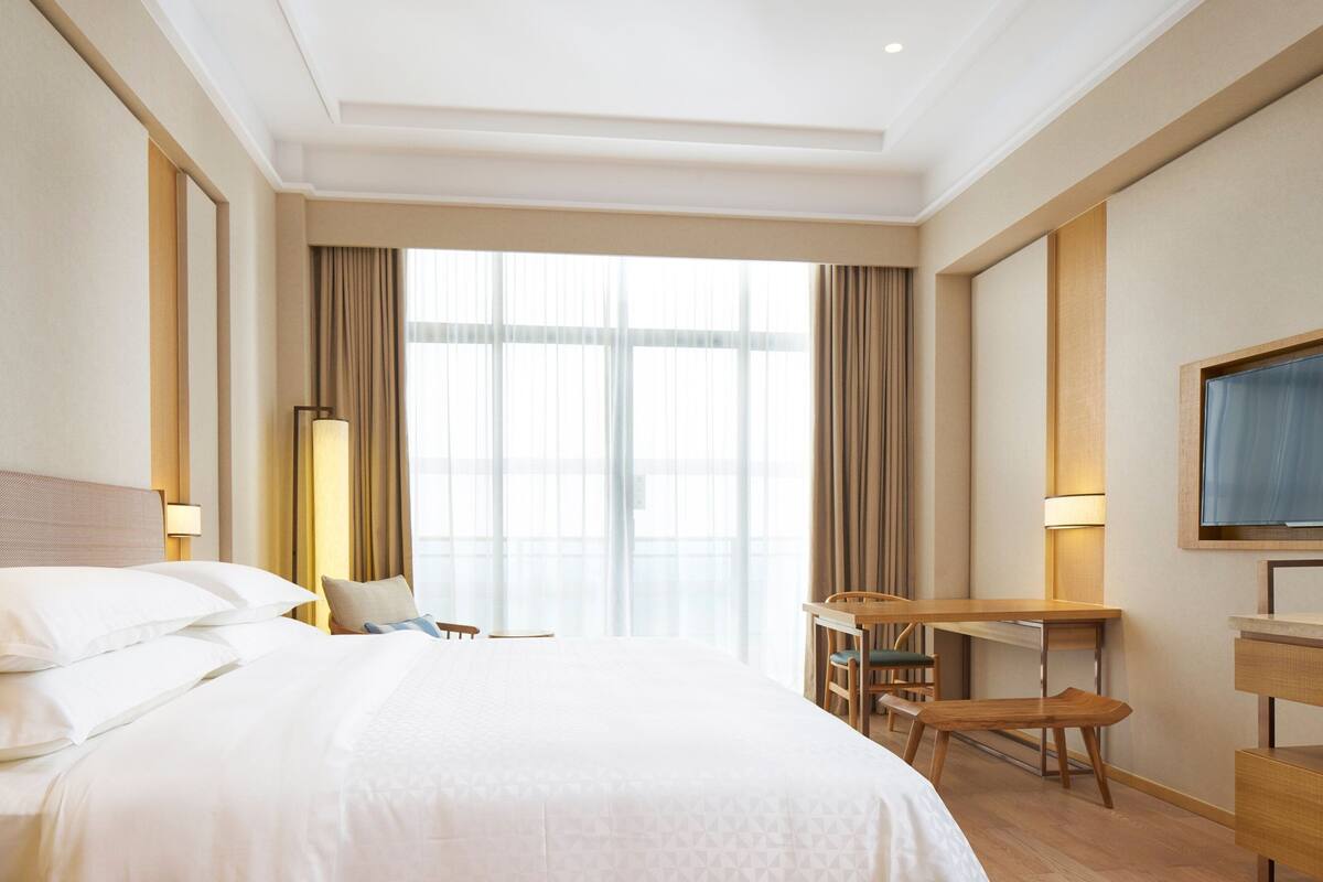 Quarto de hotel com uma cama grande, uma escrivaninha, uma cadeira, uma televisão e uma janela com cortinas.