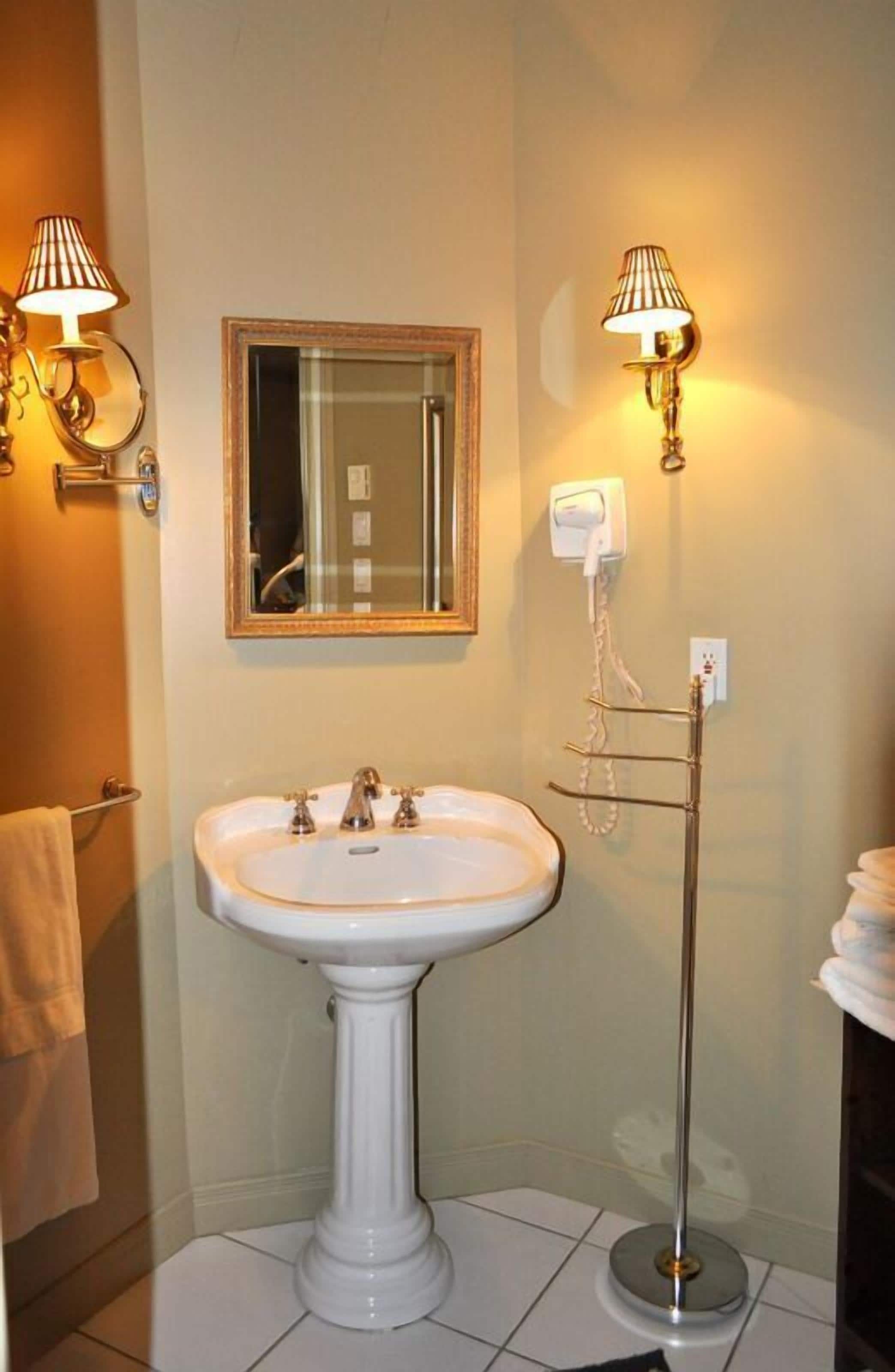 A bathroom with a pedestal sink, two wall-mounted lamps, a mirror, and a towel rack.