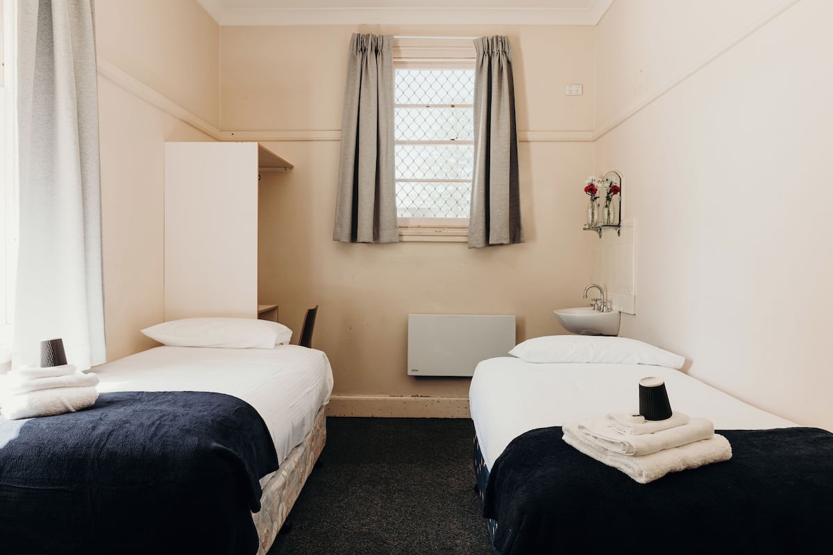 Quarto de hotel com duas camas, janela com cortinas, radiador, pia e espelho na parede.